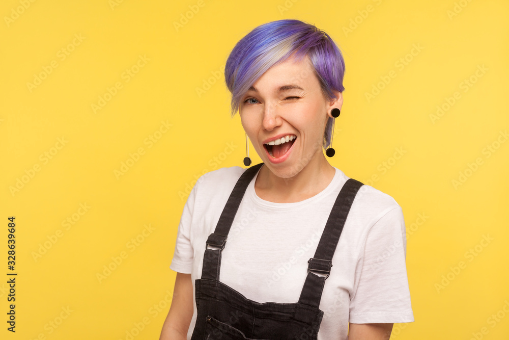 Portrait of excited cheerful hipster woman with violet dyed short hair in denim overalls winking at camera and smiling, flirting with friendly playful look. isolated on yellow background, studio shot Portrait of excited cheerful hipster woman with violet dyed short hair in denim overalls winking at camera and smiling, flirting with friendly playful look. isolated on yellow background, studio shot