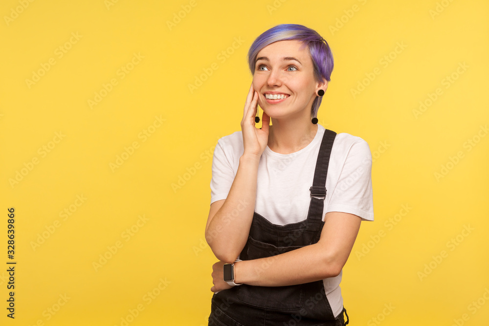 Portrait of happy beautiful hipster girl with violet short hair in denim overalls holding palm near face and dreaming pleasant fantasies, sweet memories. isolated on yellow background, studio shot Portrait of happy beautiful hipster girl with violet short hair in denim overalls holding palm near face and dreaming pleasant fantasies, sweet memories. isolated on yellow background, studio shot