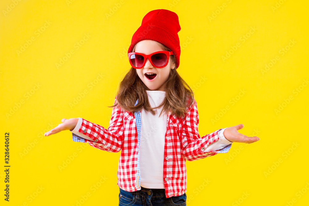Beautiful cheerful stylish little girl dressed in pink checkered shirt, red cap, sunglasses and jeans on yellow background. Surprised shocked cute child is shrugging. Emotional portrait concept. Beautiful cheerful stylish little girl dressed in pink checkered shirt, red cap, sunglasses and jeans on yellow background. Surprised shocked cute child is shrugging. Emotional portrait concept.