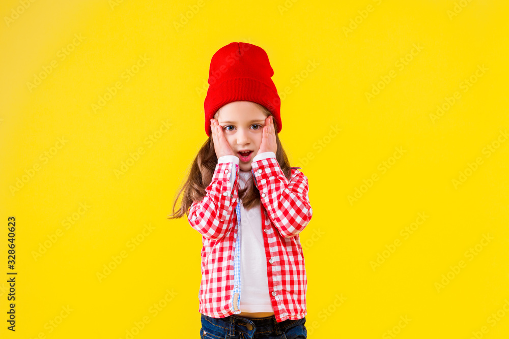 Beautiful stylish little girl dressed in pink checkered shirt, red cap and jeans on yellow orange background. Surprised shocked cute child put hand to cheek. Emotional portrait concept.