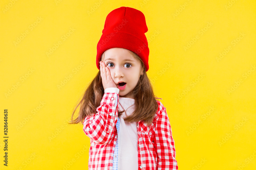 Beautiful stylish little girl dressed in pink checkered shirt, red cap and jeans on yellow orange background. Surprised shocked cute child put hand to cheek. Emotional portrait concept. Beautiful stylish little girl dressed in pink checkered shirt, red cap and jeans on yellow orange background. Surprised shocked cute child put hand to cheek. Emotional portrait concept.