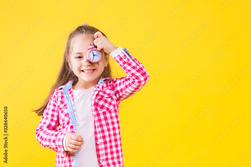 Pretty beautiful fair haired girl dressed in pink checkered shirt on yellow orange background. Cute smiling child is holding in hand in front of eye little alarm clock. Emotional portrait concept. Pretty beautiful fair haired girl dressed in pink checkered shirt on yellow orange background. Cute smiling child is holding in hand in front of eye little alarm clock. Emotional portrait concept.
