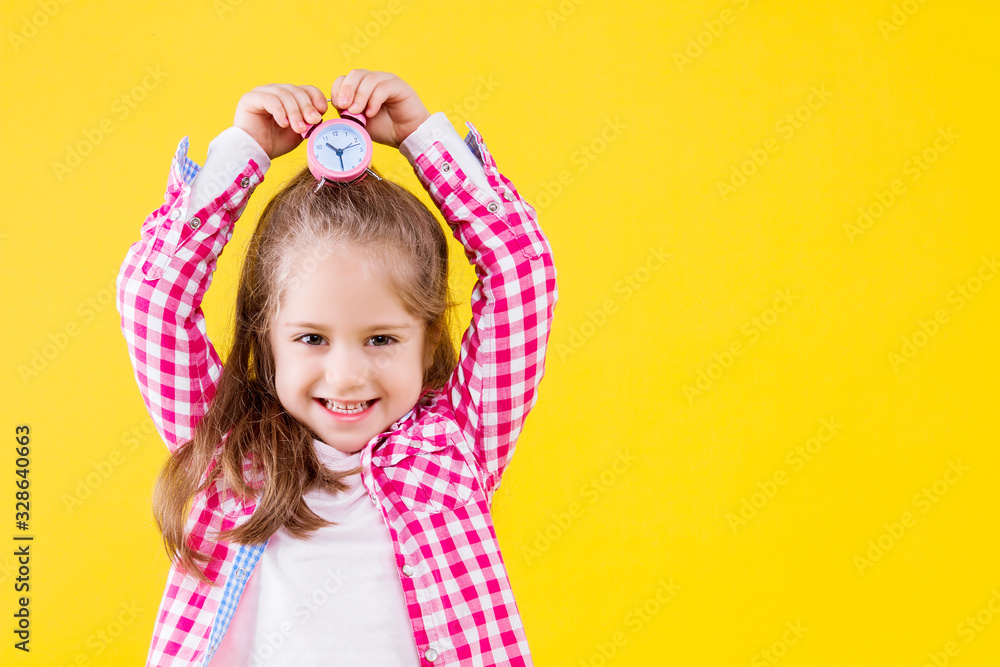 Pretty beautiful fair haired girl dressed in pink checkered shirt on yellow orange background. Cute smiling child is holding in hand little alarm clock on her head. Emotional portrait concept. Pretty beautiful fair haired girl dressed in pink checkered shirt on yellow orange background. Cute smiling child is holding in hand little alarm clock on her head. Emotional portrait concept.