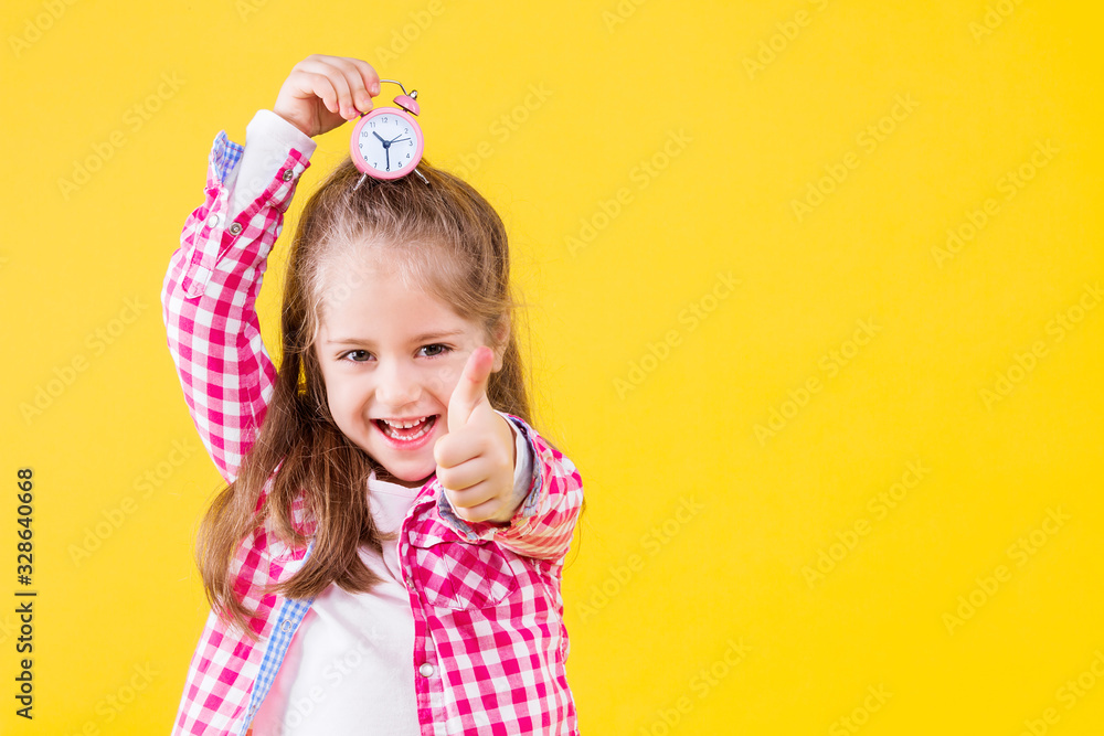 Pretty beautiful fair haired girl dressed in pink checkered shirt on yellow orange background. Cute smiling child is holding in hand little alarm clock on her head. Emotional portrait concept. Pretty beautiful fair haired girl dressed in pink checkered shirt on yellow orange background. Cute smiling child is holding in hand little alarm clock on her head. Emotional portrait concept.
