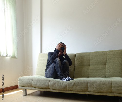 Frustrated man touching his head and keeping eyes closed while sitting on the couch at home. Feeling stressed.