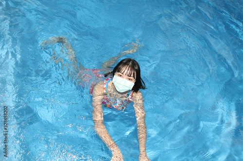 Cute little asian woman swimming in blue water pool with protective face mask