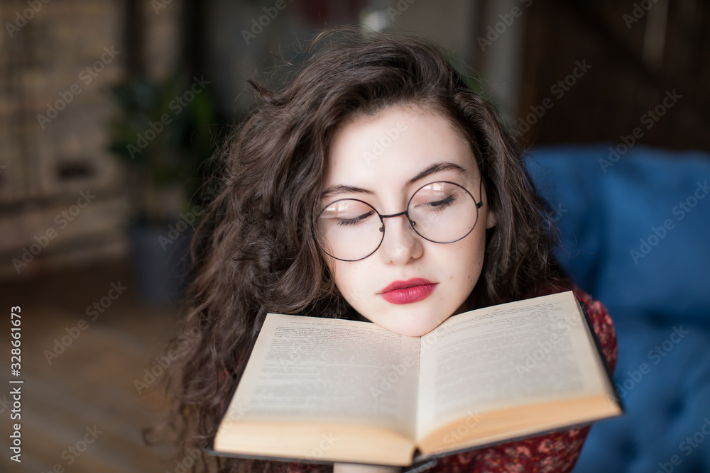 Close up portrait of attractive student girl with curly hair, in round glasses. Young woman sitting and reading book in hands near face, place her chin on it. Education and knowledge concept. Close up portrait of attractive student girl with curly hair, in round glasses. Young woman sitting and reading book in hands near face, place her chin on it. Education and knowledge concept.