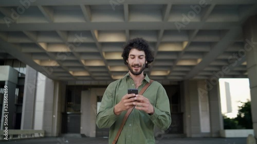 Smiling professional young executive man come out of the corporate building using his smart phone walking toward camera
