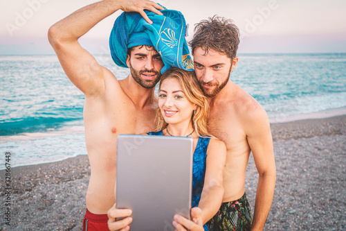 Young friends holding digital tablet in front of them on beach in summer taking a selfie together. People having fun in the summertime joking and taking pictures in front of the sea.