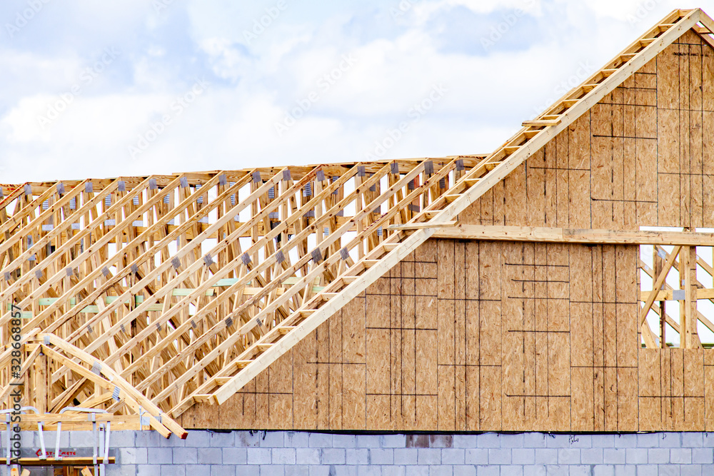 Wooden roof trusses on a new home under construction are shown on a sunny day. A second home under construction is partially shown in the background.
