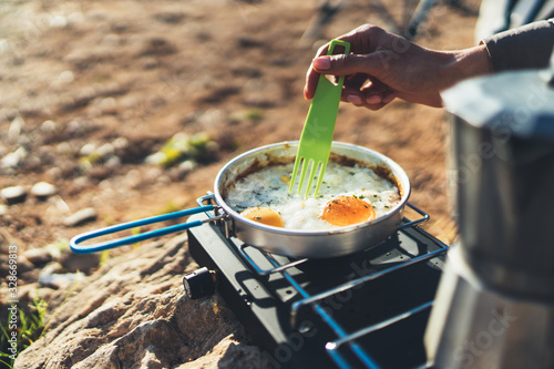 person cooking scrambled eggs in nature camping outdoor, cooker prepare breakfast picnic on metal gas stove, tourism recreation outside; campsite lifestyle