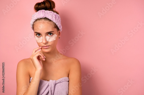 A girl with clean skin stands on a pink background in a towel and patches under her eyes and looks incredulously into the frame