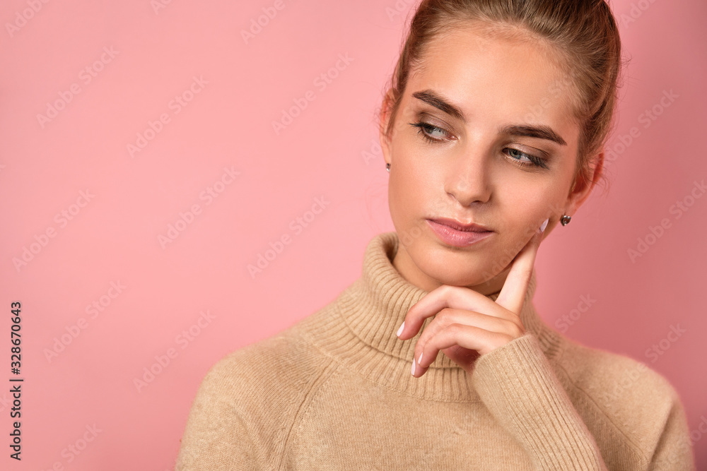 A girl with clean skin and a high bun stands on pink background and looks thoughtfully to the side, propping her cheek with finger A girl with clean skin and a high bun stands on pink background and looks thoughtfully to the side, propping her cheek with finger