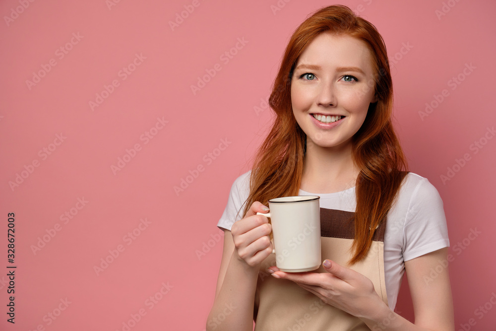The red-haired girl in an apron stands on a pink background with a white cup in her hand and smiles broadly The red-haired girl in an apron stands on a pink background with a white cup in her hand and smiles broadly