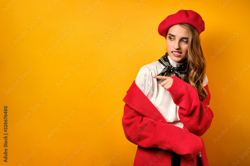 A beautiful girl in a red coat and beret stands on a yellow background with horror, looks away pointing finger there A beautiful girl in a red coat and beret stands on a yellow background with horror, looks away pointing finger there