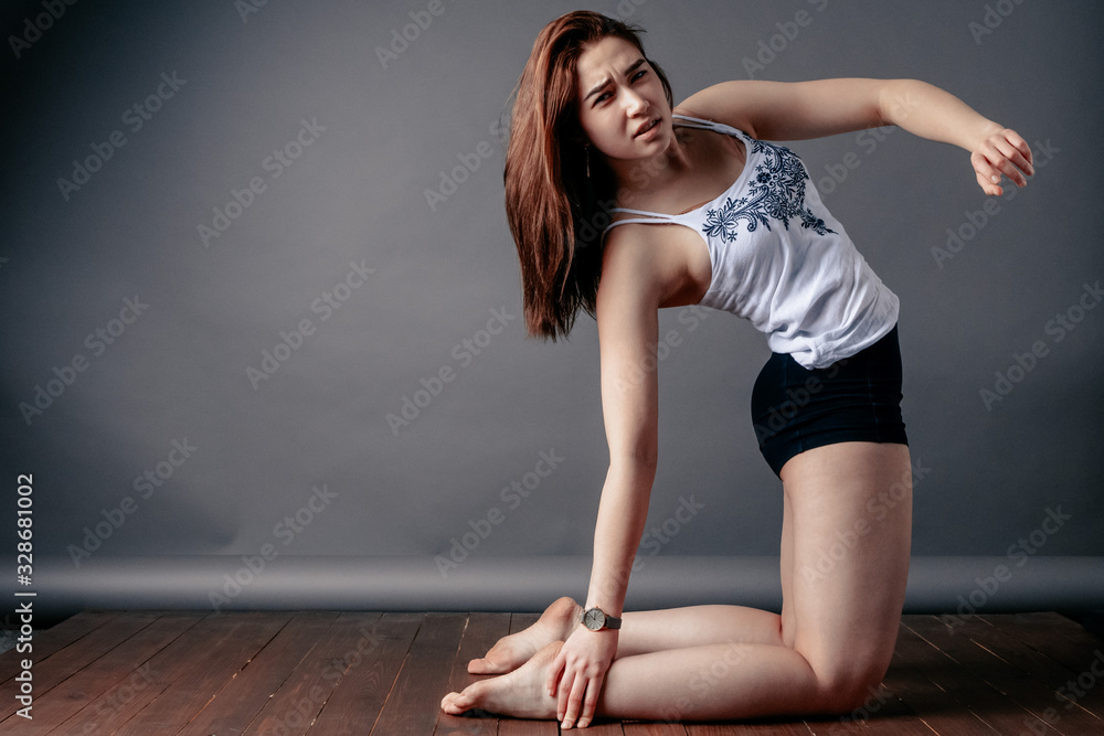 Obraz premium Beautiful young woman posing on a gray background. A woman tries to bend in the back and fails. Girl in sportswear shows exercise, beginner gymnast, displeased facial expression