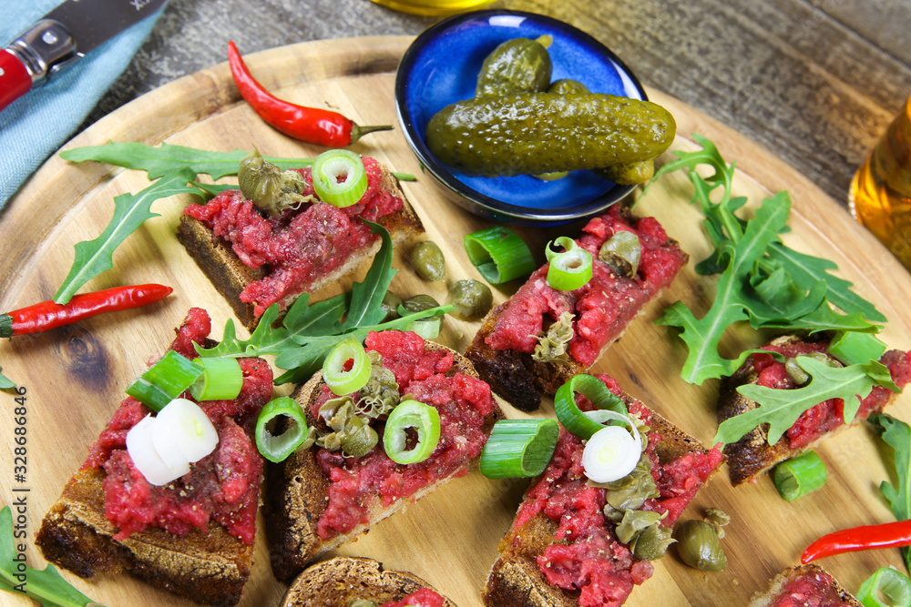 Hearty hefty german snack: isolated wood plate with roasted beef tatar garlic bread, spring onions, capers, pickled cucumbers (gherkins) and rucola salad leaves
