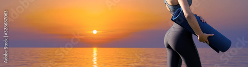 A young slim athletic girl in sportswear performs a set of exercises on the background of the sunset by the sea. Stands with a yoga mat.