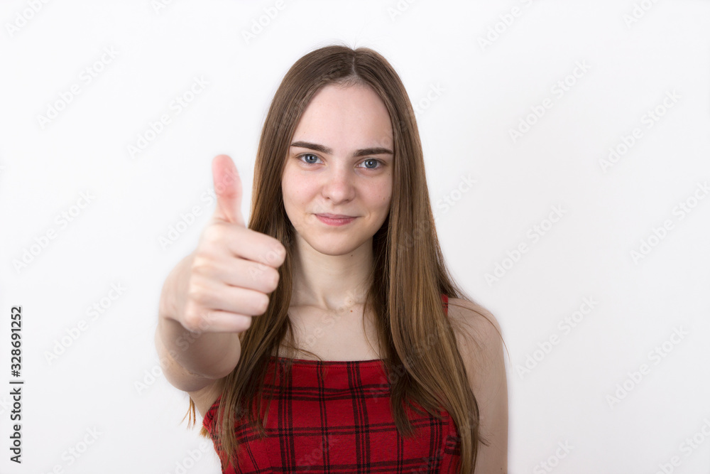 young fair-haired teenager girl with long hair, without makeup shows thumb up. Girl in a plaid sleeveless dress on white background young fair-haired teenager girl with long hair, without makeup shows thumb up. Girl in a plaid sleeveless dress on white background