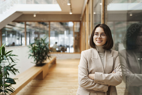 Business, education and women concept. Portrait of successful young caucasian woman in elegant jacket, leaning on glass wall in office hall waiting for coworker business center, smiling happy