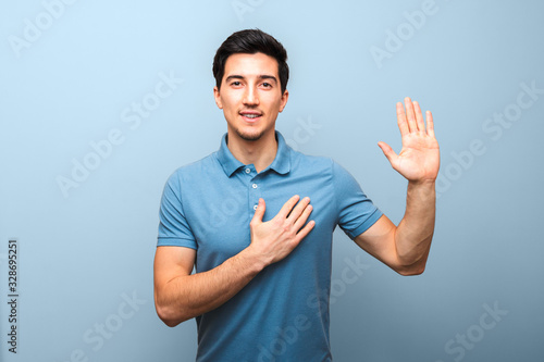 handsome young man with neutral smile in blue polo shirt with hand on chest giving oath against blue background. studio shoot photo