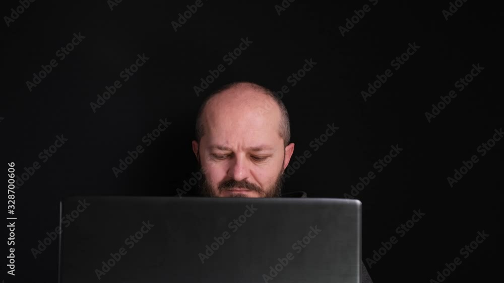 Man working from home office. Caucasian man in hoodie sitting behind the laptop and working at home, putting headphones, black background Man working from home office. Caucasian man in hoodie sitting behind the laptop and working at home, putting headphones, black background
