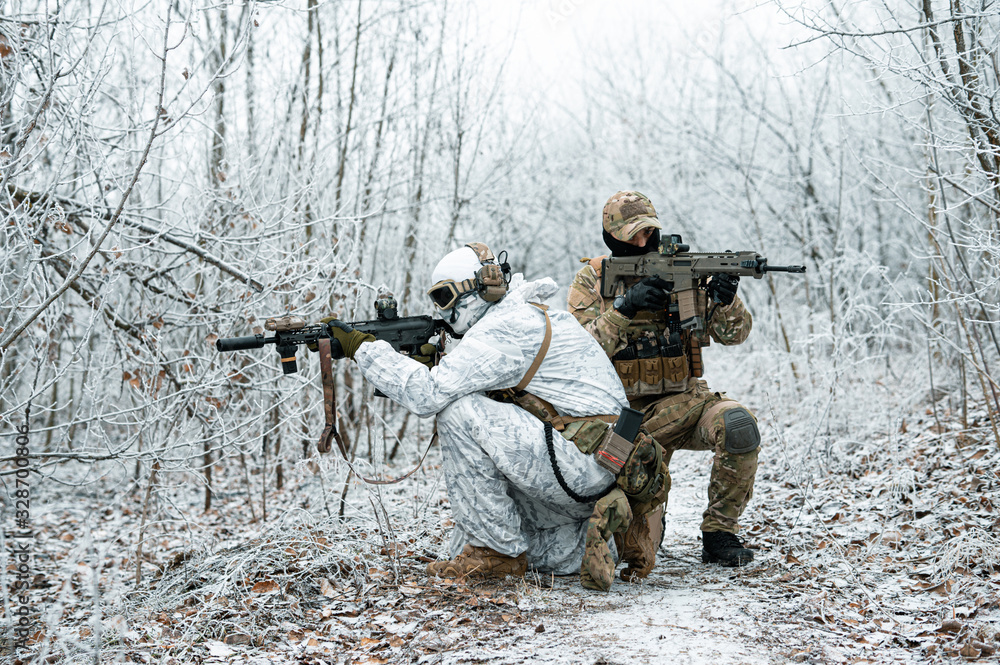 Fototapeta premium Two men in camouflage uniform with machineguns back to back. Soldiers with muchinegun stood on knelt in the winter forest. Side view.