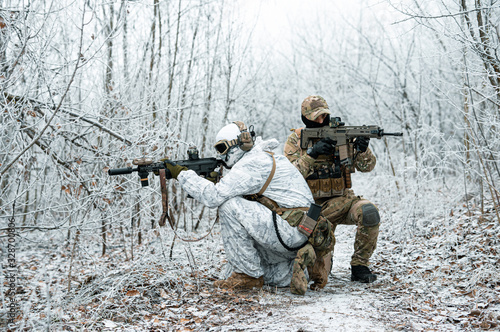 Two men in camouflage uniform with machineguns back to back. Soldiers with muchinegun stood on knelt in the winter forest. Side view.