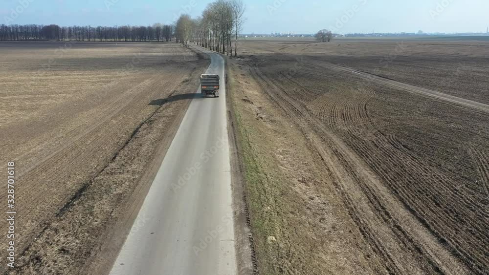 The truck moves fast along a country road. The village road. View from above. Alone Dirty Truck driving on clear spring asphalt road against blue sky with white clouds. The truck moves fast along a country road. The village road. View from above. Alone Dirty Truck driving on clear spring asphalt road against blue sky with white clouds.