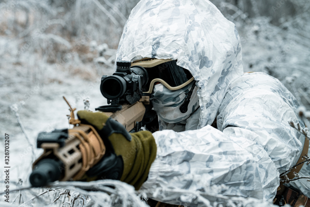 Closeup military man in white camouflage uniform with hood and machinegun in the long winter grass. Soldier stood on knelt with machinegun.