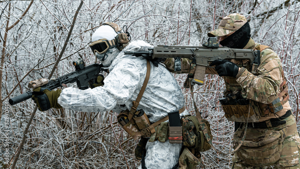 Fototapeta premium Two men in camouflage white, green uniform with machineguns. Soldiers with muchineguns go one after another in the winter forest. Horizontal photo