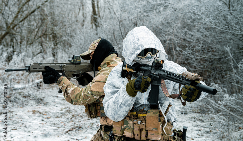 Fototapeta premium Two men in camouflage white, green uniform with machineguns back to back. Soldiers with muchinegun in the winter forest. Horizontal photo, side view.