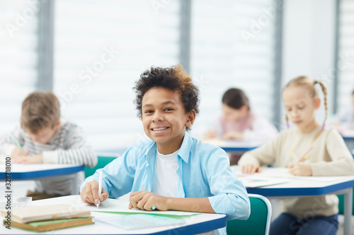 Horizontal medium close up portrait of happy mixed-race boy with kinky hair sitting at school desk looking at camera smiling, copy space