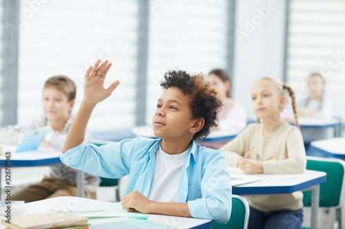 Horizontal portrait of smart middle school male student raising his hand to give answer in class, copy space
