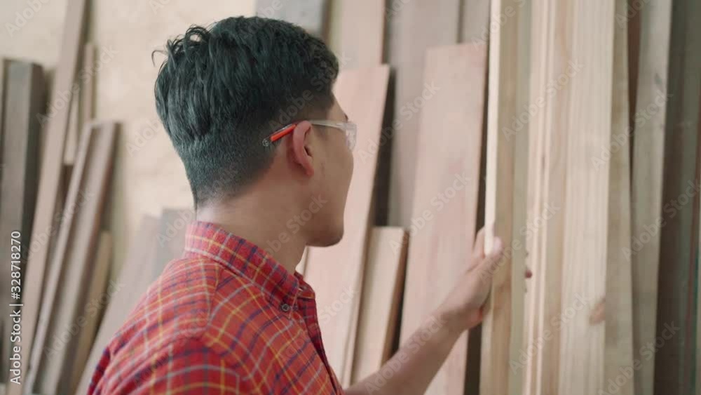 Carpenter working in a carpentry shop. Young asian woodworker choosing some wood for make a furniture such as modern chair. Young artisan craftsman entrepreneur. Medium to close up shot. Carpenter working in a carpentry shop. Young asian woodworker choosing some wood for make a furniture such as modern chair. Young artisan craftsman entrepreneur. Medium to close up shot.