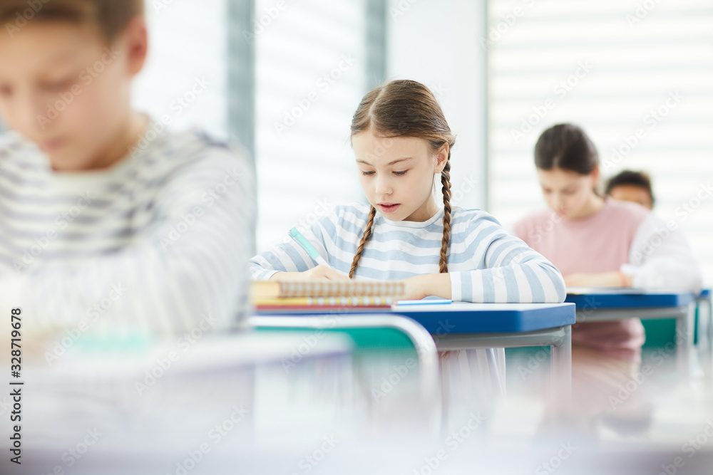 Young Caucasian girl with brown hair wearing striped seatshirt sitting at school desk in modern clasroom writing something in her notebook, copy space Young Caucasian girl with brown hair wearing striped seatshirt sitting at school desk in modern clasroom writing something in her notebook, copy space