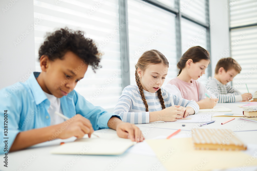 Group of four middle school students sitting together at desk in modern classrooms completing task in their notebooks, copy space Group of four middle school students sitting together at desk in modern classrooms completing task in their notebooks, copy space