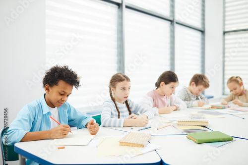 Group of five middle school students wearing casual outfits sitting at desk in modern classroom completing task, horizontal shot, copy space photo