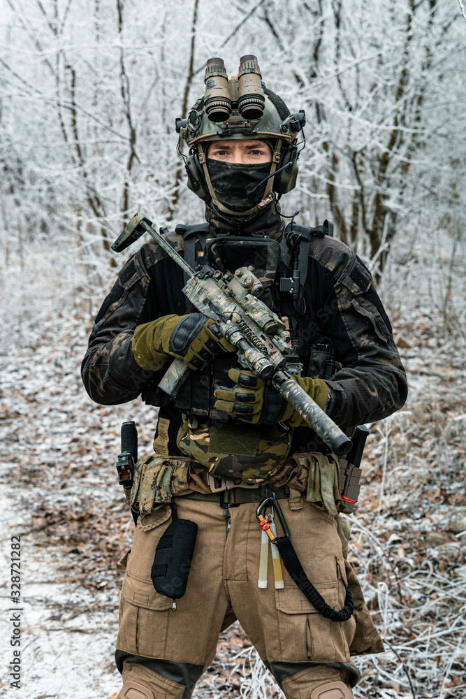 Fototapeta premium Airsoft man in black camouflage uniform and short machinegun with optical sight. Soldier in the winter forest. Vertical photo front view.