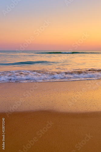 Colorful sunset at the tropical beach, sun behind clouds reflects on water and waves with foam hitting sand.