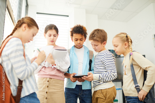 Group of five middle schoolers standing together in school corridor searching information about their homework project task during break, horizontal shot photo