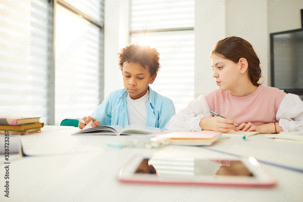 African American boy and Caucasian girl sitting together at school desk in modern classroom, girl looking at boy's textbook, copy space African American boy and Caucasian girl sitting together at school desk in modern classroom, girl looking at boy's textbook, copy space