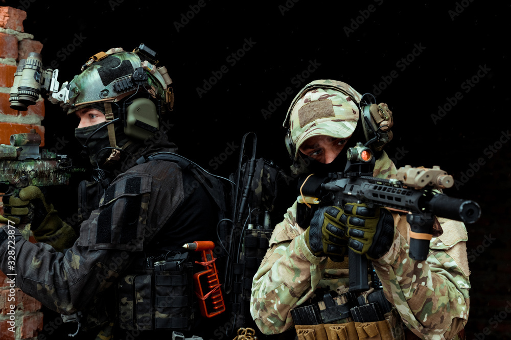 Fototapeta premium Closeup two men in camouflage cloth and black uniform with machineguns stand beside brick wall. Soldiers with muchinegun aims aiming