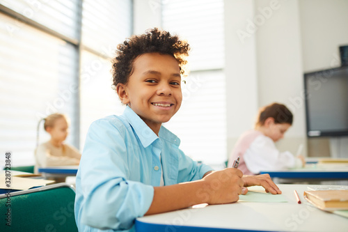 Horizontal low angle medium close up portrait of happy mixed-race boy with kinky hair sitting at school desk looking at camera smiling, copy space photo