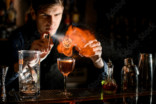 Young bartender professionally sprinkles burning slice of citrus for cocktail.