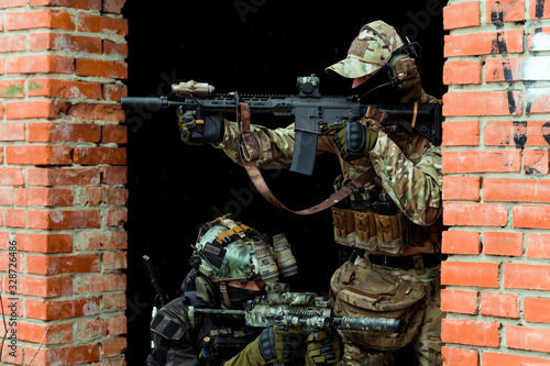 Two men in camouflage cloth green and black uniform with machineguns. Soldiers with muchinegun aims aiming in oposite sides, standing beside wall