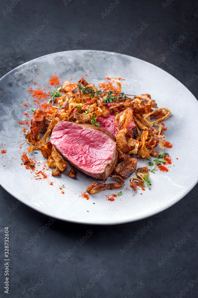 Barbecue dry aged beef fillet medallion steak natural with fried onion rings and carrots as closeup on a modern design plate with copy space Barbecue dry aged beef fillet medallion steak natural with fried onion rings and carrots as closeup on a modern design plate with copy space