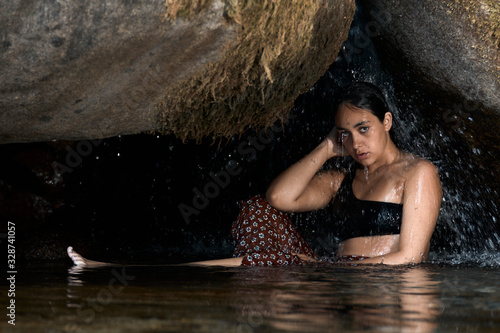 Joven morena de pelo bajo la cascada en una cueva en las montañas al aire libre.