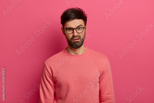 Discontent young bearded man has disgusted displeased expression, reacts on something unpleasant, frowns face, wears spectacles and jumper, stands indoor against pink background. Emotions concept