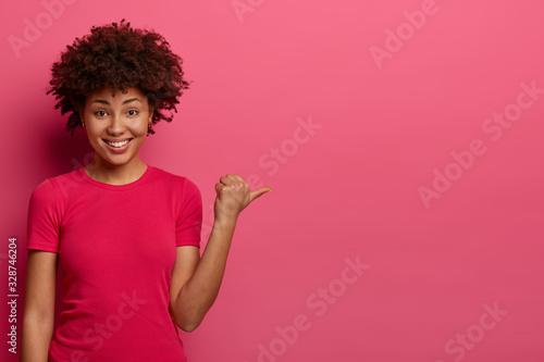 Positive young woman shop assistant helps customer to find dressing room, advertises product on sale, points thumb aside, wears casual t shirt, smiles gladfully, isolated on pink background.